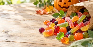 Colorful gummy candies, fruit jellies, sour sugar-coated strips and hard candies spilling from a kraft paper bag onto a wooden table, with a carved pumpkin and autumn leaves in the background.